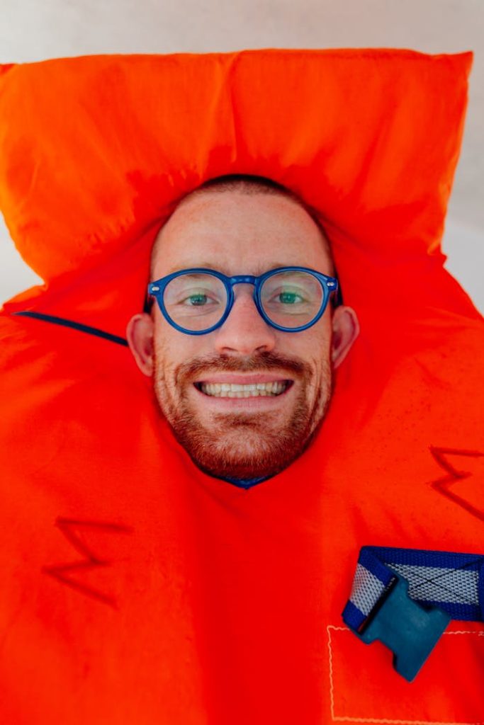 Close-up portrait of a happy man wearing glasses and a bright orange life vest, exuding humor.